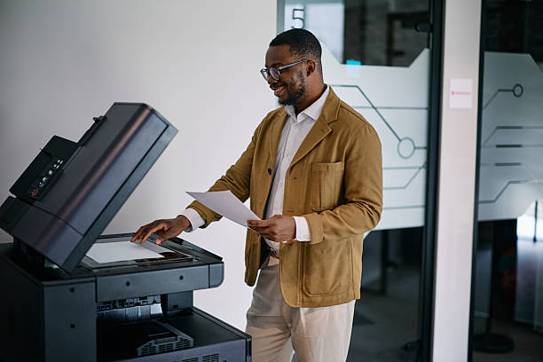 Smiling mid adult african businessman making a copy using a modern multifunction printer in the office, he is holding a paper document and placing another one on the scanner glass