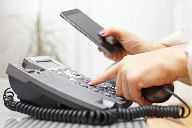 Woman is dialing on land line phone and looking on mobile phone