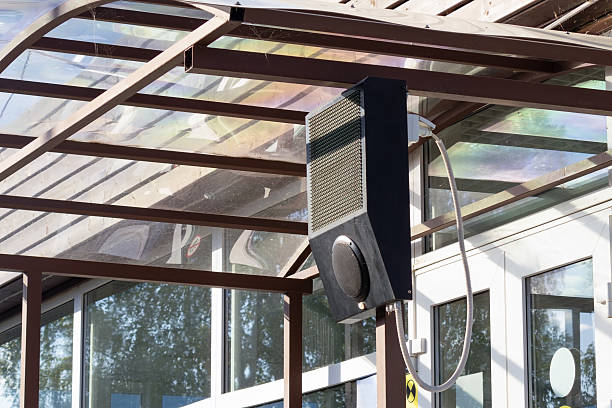Close-up view of exterior intercom with mesh grille and circular speaker mounted beneath a glass-and-metal canopy near building doors, reflecting trees and sky and forming a clean contemporary architectural detail.
