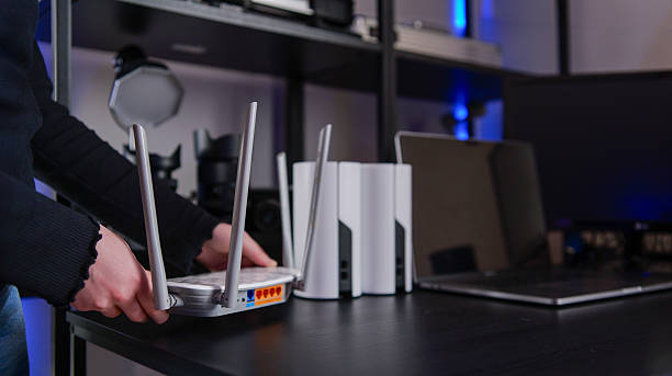 A person's hands are seen adjusting the antennas of a white wireless router, with other mesh network devices and a laptop on a dark desk
