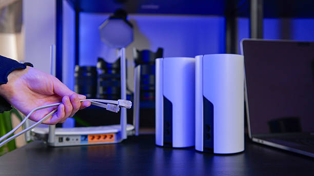 A hand connects Ethernet cables to a router, with mesh Wi-Fi units and a laptop nearby, suggesting network setup and expansion