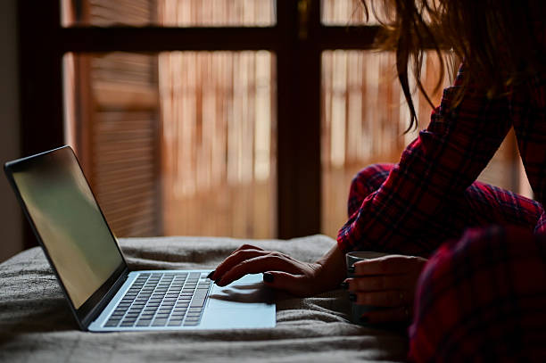 Woman typing on laptop in bed wearing pajamas, enjoying a warm cup of coffee